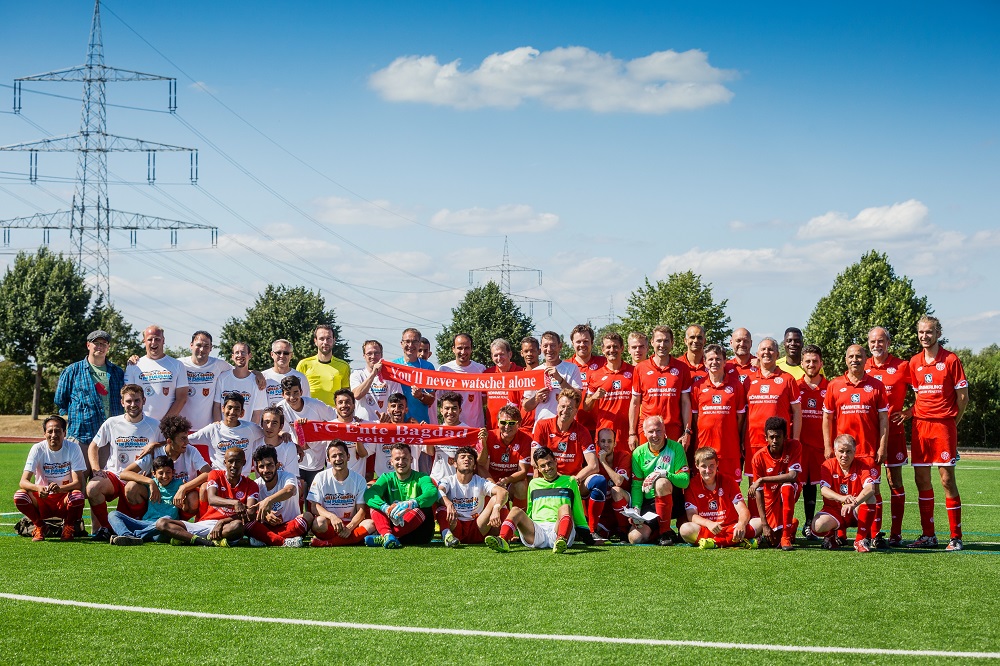Eine große Gruppe von Menschen, hauptsächlich Männer, in Fußballbekleidung steht und hockt auf einem grünen Fußballfeld. Viele tragen rote Trikots, einige blau-weiße. Im Hintergrund ist ein Mast mit der Aufschrift "You'll never walk alone" zu sehen. Im Vordergrund hält eine Gruppe von Kindern ein Banner mit der Aufschrift "FC Ente Baghdad seit 1972".