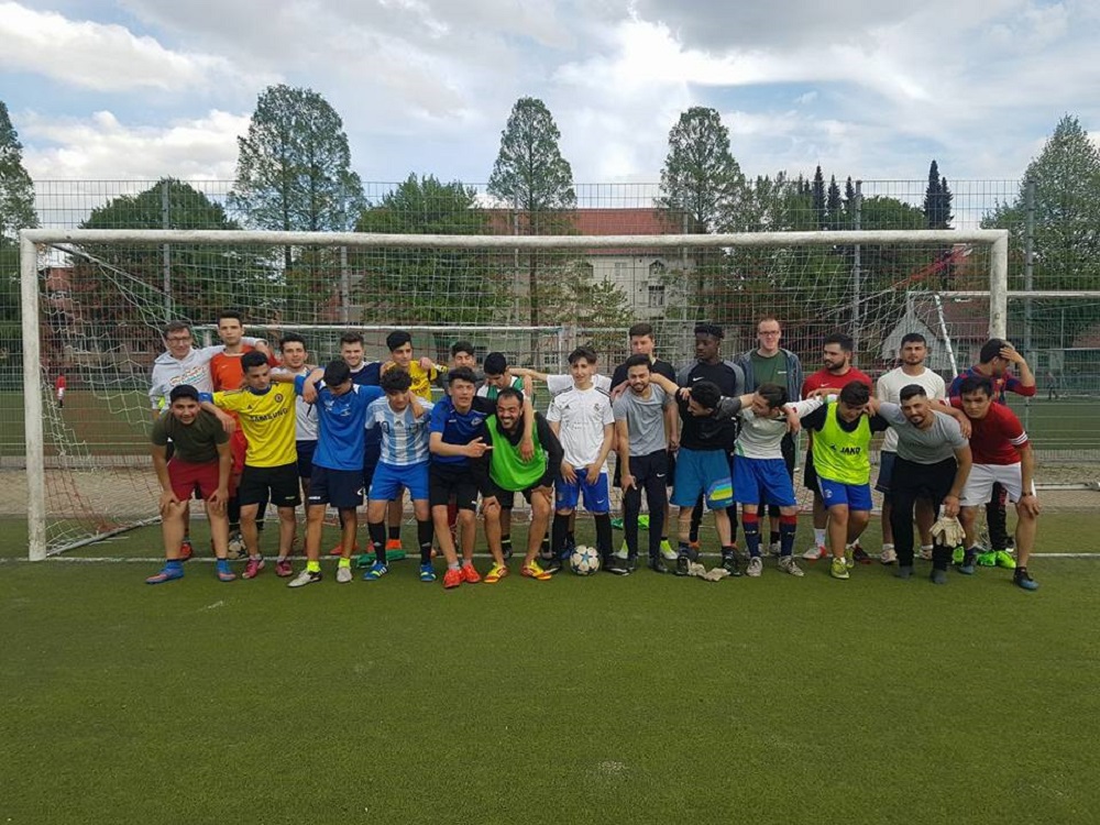 Eine große Gruppe von Jugendlichen und jungen Erwachsenen steht auf einem Fußballfeld vor einem Fußballtor. Viele tragen Trainingskleidung und schauen in die Kamera. Im Hintergrund sind Bäume und Gebäude zu sehen.
