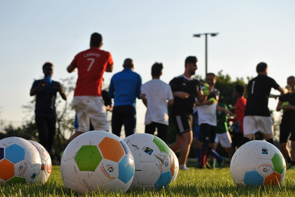 Fußbälle auf einem grünen Rasen vor einer Gruppe von Menschen, die in einem Kreis stehen. Im Hintergrund ist ein Fußballfeld und ein Mast mit Flutlichtern zu sehen.