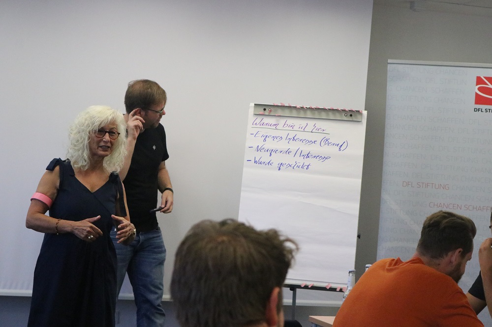 Eine Frau mit gelockten, grauen Haaren steht vor einer Wandtafel und spricht. Um sie herum sitzen mehrere Männer, vermutlich Teilnehmer einer Trainerschulung. Auf der Tafel sind Stichpunkte zum Thema Jugendförderung und Trainerstärken notiert. Im Hintergrund ist ein Logo der DFL Stiftung zu sehen.