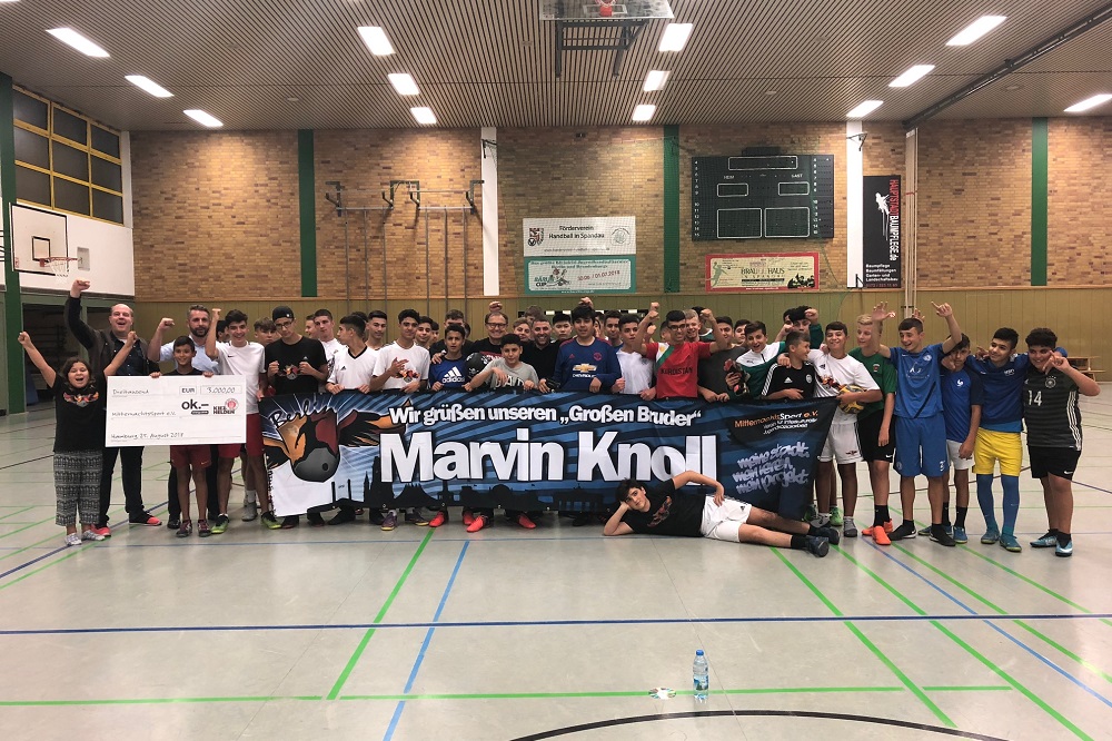 Eine große Gruppe von Jugendlichen und Erwachsenen steht in einer Turnhalle zusammen und hält einen großen Scheck mit der Aufschrift "Marvin Knoll" hoch. Im Hintergrund sind Banner und Plakate zu sehen. Die Stimmung ist fröhlich und gemeinschaftlich.
