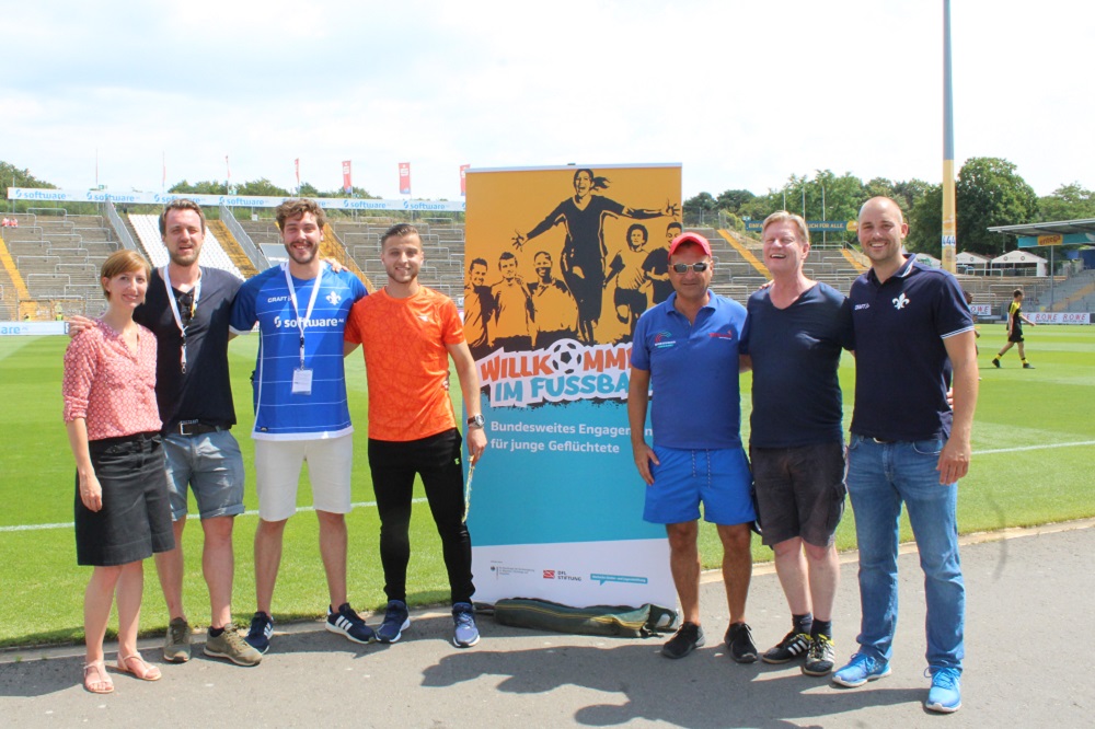 Eine Gruppe von sieben Personen steht auf einem Fußballfeld vor einem Banner mit der Aufschrift "Willkommen im Fußball - Bundesweites Engagement für junge Geflüchtete". Im Hintergrund ist ein Fußballstadion zu sehen.