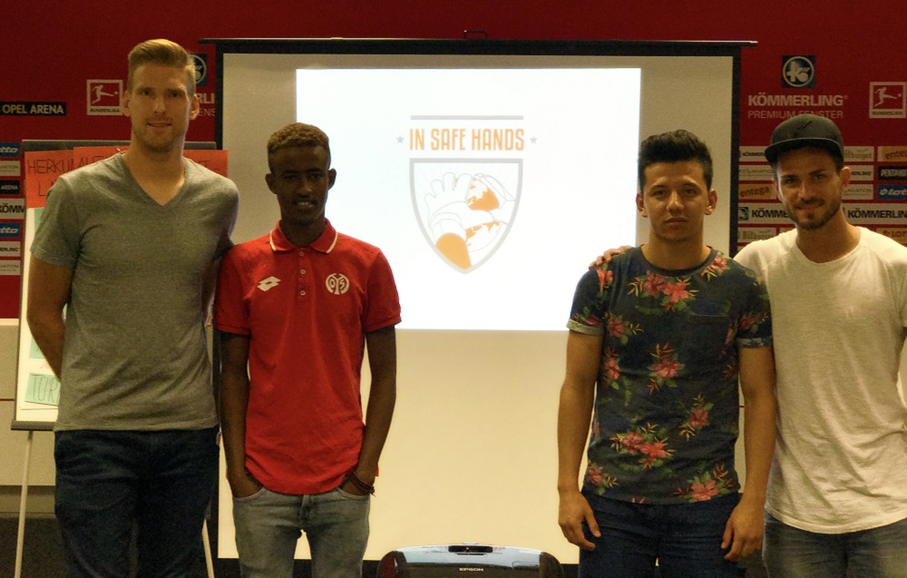 Vier junge Menschen stehen vor einer Projektionswand mit dem Logo "In safe hands e.V." und einem Fußball. Der Spieler Danny Latza steht links im Bild.