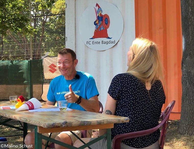 Zwei Personen sitzen an einem Tisch im Freien und unterhalten sich. Der Mann trägt ein blaues T-Shirt mit der Aufschrift "Wir lieben Fußball!". Im Hintergrund ist ein Banner mit dem Logo "FC Ente Bagdad" zu sehen.
