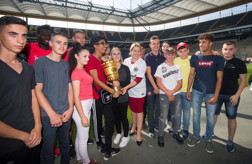 Eine Gruppe Jugendlicher steht im Stadion mit der Bundesliga-Meisterschale. Einige tragen T-Shirts von Eintracht Frankfurt und Adidas. Im Hintergrund sind die Sitze und die Struktur des Stadions zu sehen.