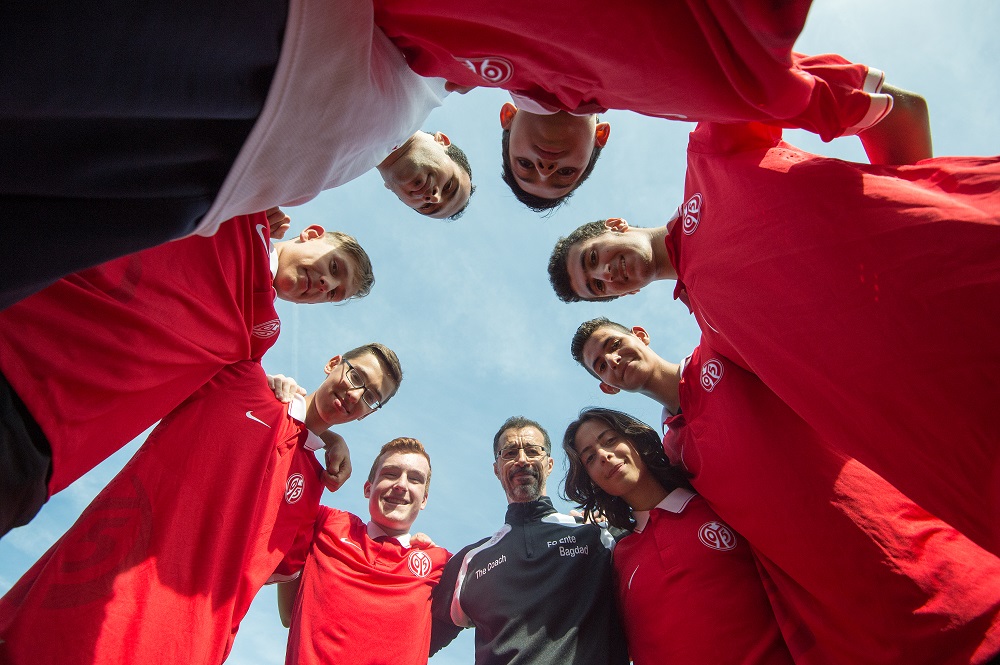 Eine Gruppe junger Fußballspieler im roten Trikot steht im Kreis und blickt nach oben. Im Zentrum steht ein Mann mit Trainingsanzug, vermutlich der Trainer. Der blaue Himmel ist im Hintergrund zu sehen.