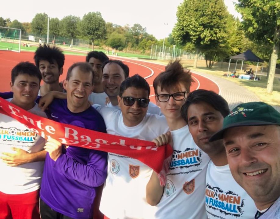 Eine Gruppe von Männern steht lächelnd auf einem Fußballfeld. Sie tragen rote Trikots und einige haben eine rote Fahne mit der Aufschrift "Benefiz-Fußballturnier Hechtsheim" umgelegt. Im Hintergrund ist eine Laufbahn und eine Sporthalle zu sehen.