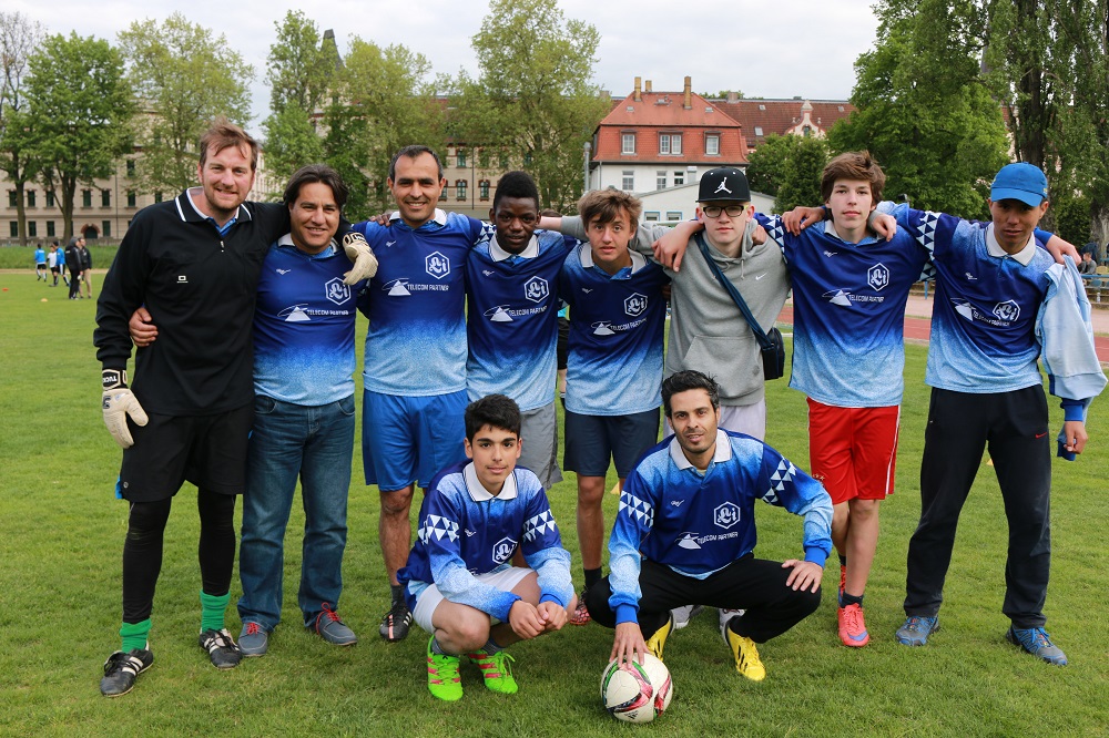 Eine Gruppe Jugendlicher in blauen Fußballtrikots mit dem Logo der DFL Stiftung posiert mit ihrem Trainer auf einem Fußballfeld. Sie lächeln in die Kamera und stehen eng zusammen. Im Hintergrund ist ein Gebäude und grüne Vegetation zu sehen.