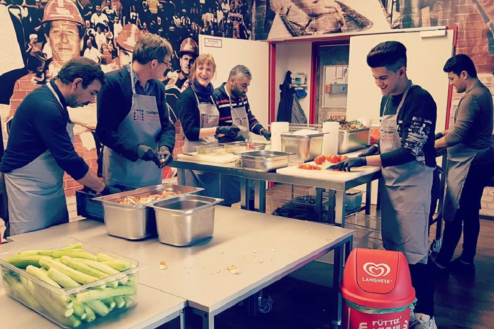 Eine Gruppe von Menschen steht um einen Tisch und bereitet Gemüse zu. Im Hintergrund ist ein Raum mit Bildern an der Wand zu sehen. Ein Mann mit schwarzem T-Shirt und Schürze schneidet Gurken. Auf dem Tisch stehen verschiedene Schüsseln mit geschnittenem Gemüse. Im Hintergrund befindet sich ein Eimer mit dem Logo von Langnese.