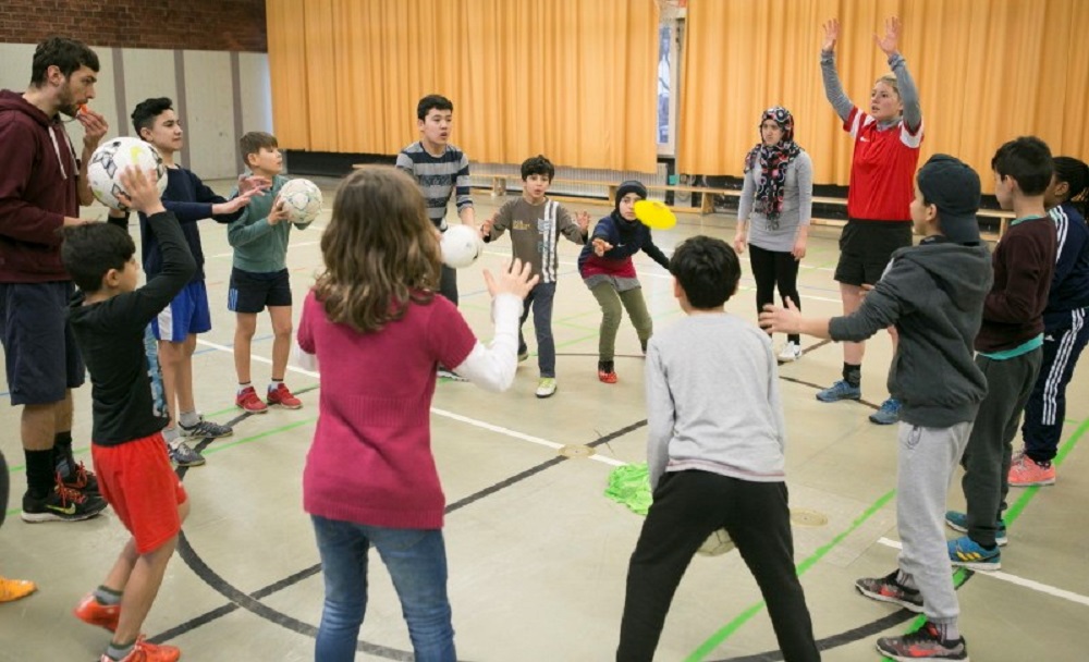 Eine Gruppe von Kindern und Jugendlichen steht in einem Turnsaal im Kreis und übt mit Bällen. Im Hintergrund sind zwei Erwachsene erkennbar, eine Frau in roter Sportbekleidung und ein Mann. Der Fokus liegt auf der spielerischen Interaktion und Bewegung der Kinder mit den Bällen.