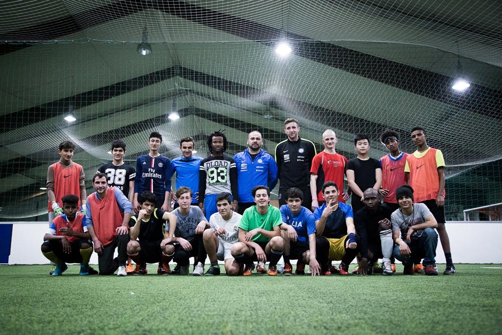 Eine Gruppe Jugendlicher in Fußballkleidung posiert auf einem grünen Kunstrasenfeld in einer Halle. In der Mitte steht Murat Korkmaz, ein Mann mit dunklen Haaren und Bart, umgeben von den Jugendlichen. Im Hintergrund befindet sich die große Halle mit einer modernen Beleuchtung.