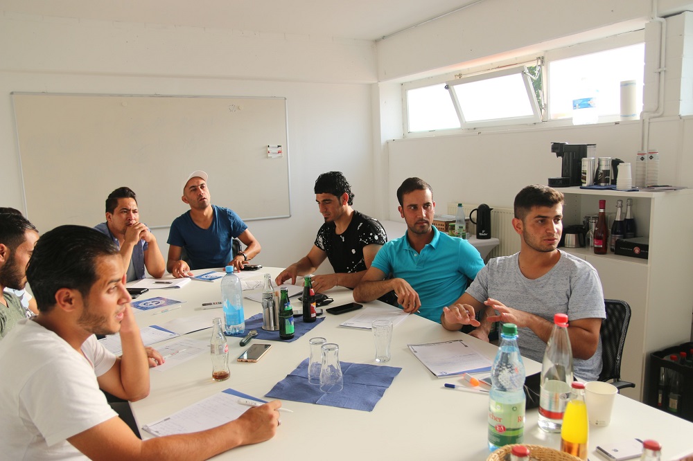 Eine Gruppe junger Männer sitzt an einem großen Konferenztisch in einem hellen Raum zusammen. Sie nehmen an einem Berufscoaching teil und wirken aufmerksam. Auf dem Tisch stehen Wasserflaschen, Kaffeetassen und weiteres Zubehör.