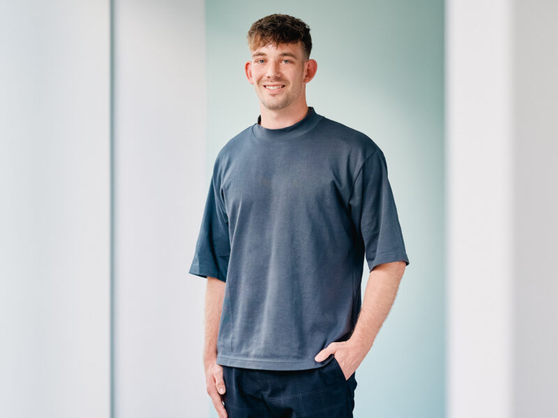 Lukas Bendix, lächelnder junger Mann mit kurzgeschorenem Haar, trägt ein dunkelblaues T-Shirt und eine dunkle Hose. Er steht vor einem hellblauen Hintergrund und hat die Hände in den Hosentaschen.