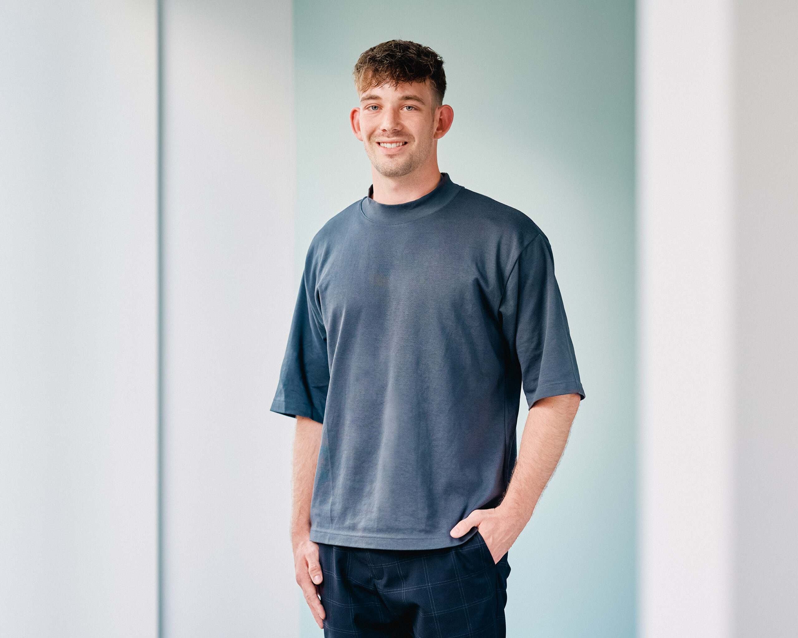 Lukas Bendix, lächelnder junger Mann mit kurzgeschorenem Haar, trägt ein dunkelblaues T-Shirt und eine dunkle Hose. Er steht vor einem hellblauen Hintergrund und hat die Hände in den Hosentaschen.