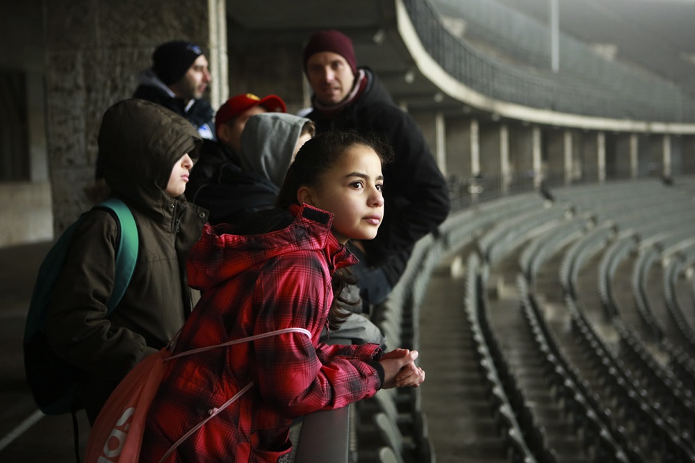 Eine Gruppe Kinder steht auf den Zuschauertribünen des Olympiastadions Berlin. Ein Mädchen im roten Winterjackett blickt konzentriert nach vorne, während die anderen Kinder und Betreuer im Hintergrund stehen. Das Bild stammt von einem Workshop der DFL Stiftung im Rahmen des Projekts "Lernort Stadion".