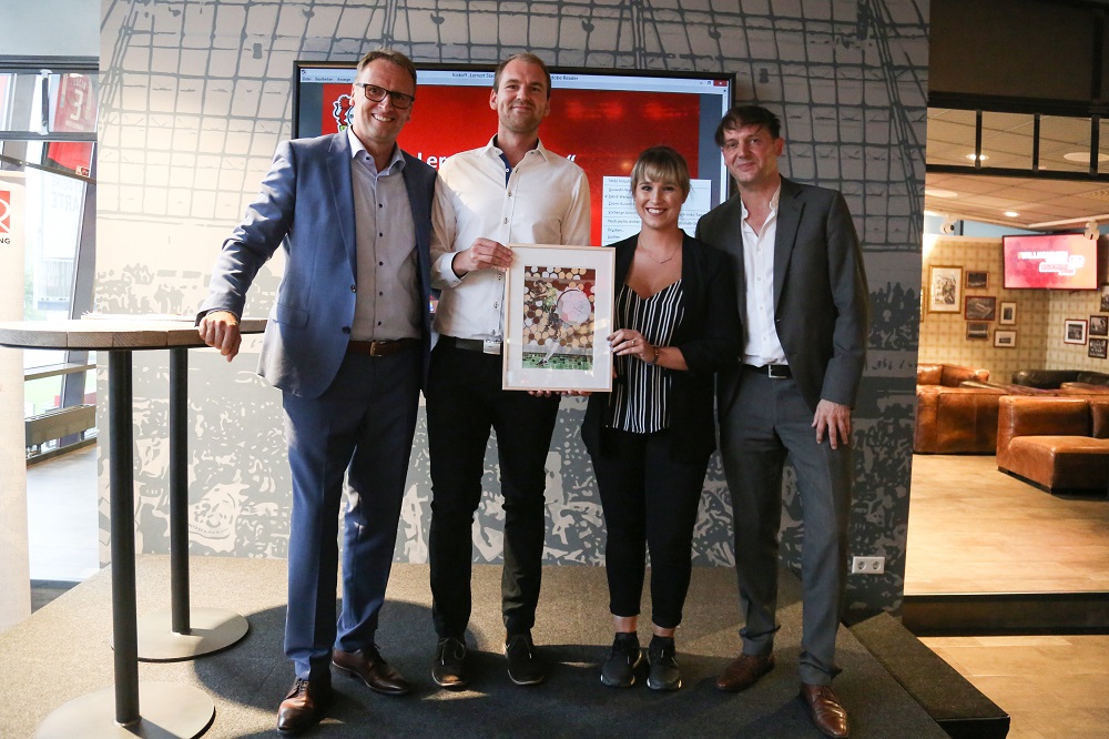 Fünf Personen stehen in einem Innenraum, wahrscheinlich in einer Lounge oder einem Büro, und halten ein gerahmtes Foto hoch. Die Personen sind gut gekleidet und lächeln in die Kamera. Im Hintergrund sind weitere Möbel und ein Bildschirm zu sehen. Das Bild zeigt vermutlich einen neuen Partner oder eine neue Initiative im Lernort Stadion.
