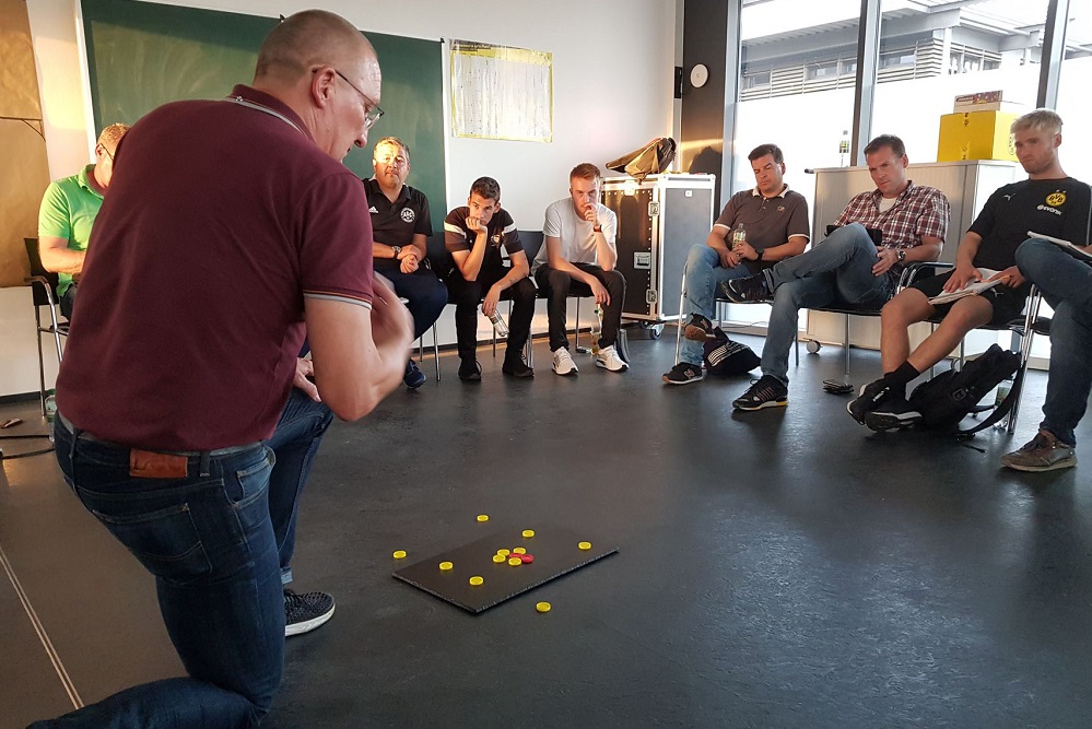 Eine Gruppe von Männern sitzt in einem Kreis auf schwarzen Stühlen in einem hellen Raum. Ein Mann steht vor der Gruppe, den Rücken zugewandt, und hält eine Hand in Richtung eines quadratischen, schwarzen Bretts mit bunten Markern darauf. Es scheint eine Übung oder ein Workshop im Rahmen der Trainer-Ausbildung der DFL Stiftung zu sein.