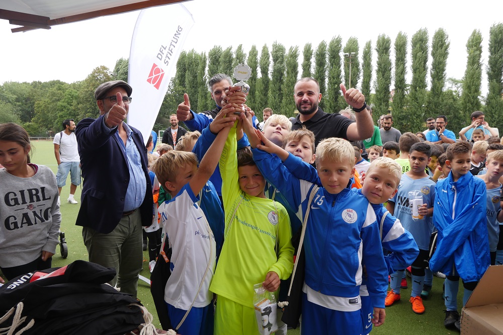 Eine Gruppe von Kindern in Fußballtrikots steht auf einem grünen Rasen und hebt gemeinsam einen Pokal in die Höhe. Im Hintergrund sind weitere Personen und ein Banner mit der Aufschrift "DFL Stiftung" zu sehen. Die Kinder strahlen und zeigen Freude über den Gewinn.