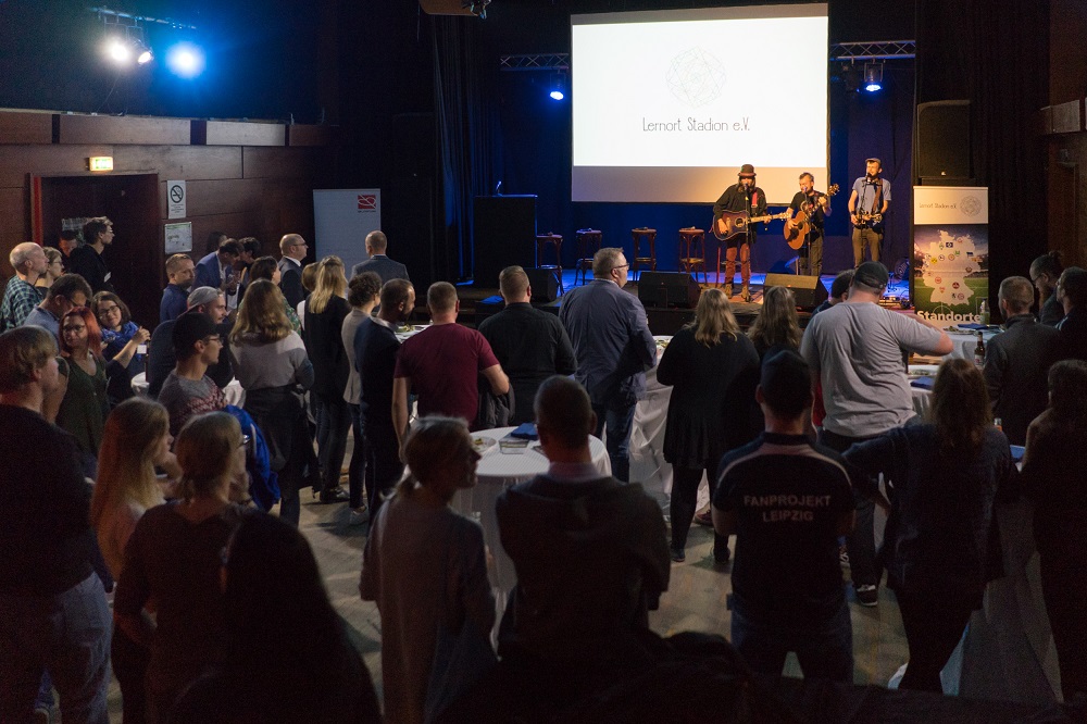 Eine große Menschenmenge nimmt an einer Veranstaltung im Innenraum einer Halle teil. Im Hintergrund ist eine Bühne mit zwei Musikern zu sehen, die Gitarren spielen. Auf einer Leinwand im Hintergrund steht "Lernort Stadion e.V.". Die Veranstaltung scheint ein Treffen des Lernort Stadion e.V. zu sein, möglicherweise ein öffentlicher Abend oder eine Präsentation.