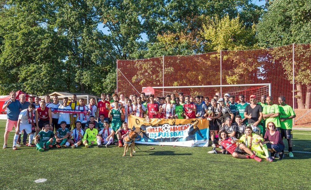 Eine große Gruppe von Kindern und Jugendlichen steht auf einem Fußballfeld in Nürnberg zusammen und lächelt in die Kamera. Sie tragen verschiedene Fußballtrikots und Sportkleidung. Im Hintergrund ist ein Fußballtor zu sehen. Vor der Gruppe wird ein Banner mit der Aufschrift "Willkommen im Fußball" gehalten.