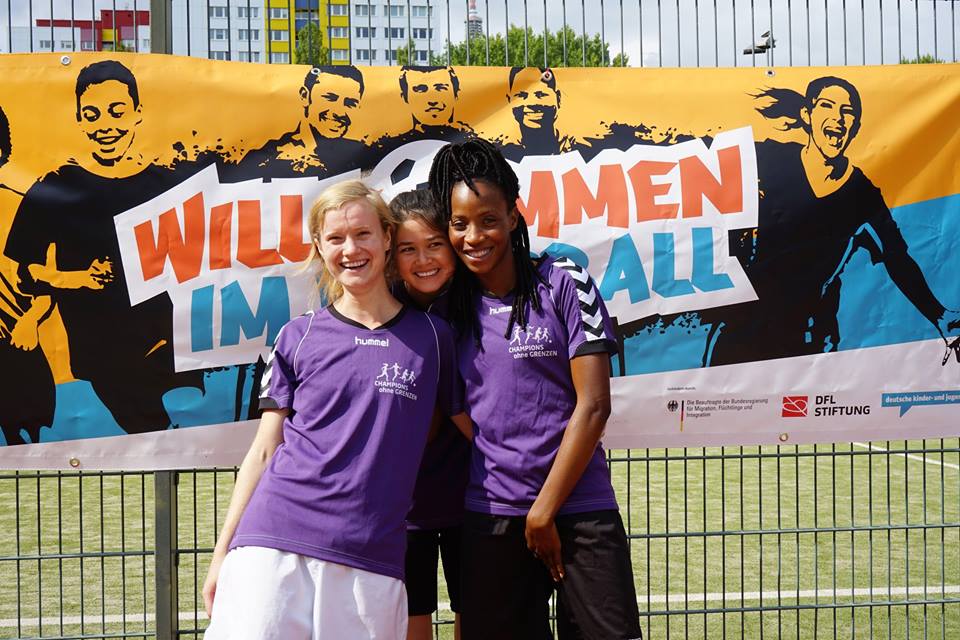 Drei lächelnde Frauen stehen vor einer großen Bannerwand mit der Aufschrift "WIR KICKEN GEGEN RASSISMUS" und dem Logo der DFL Stiftung. Im Hintergrund ist ein Fußballfeld erkennbar.