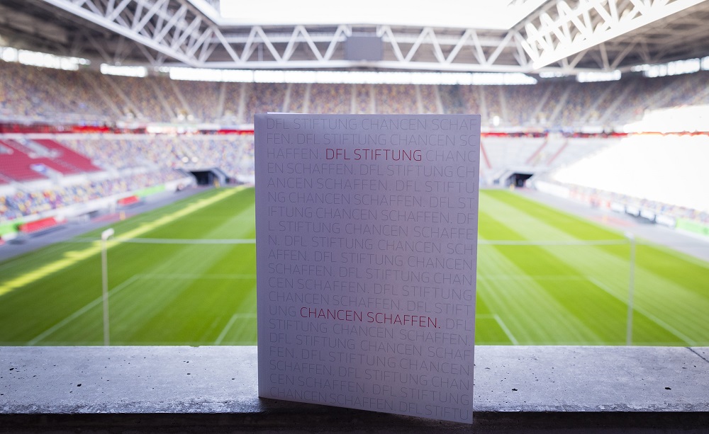 Plakat mit der Aufschrift "SCHAFFEN. DFL STIFTUNG Chancen" steht vor verschwommenem Fußballstadion, Fokus auf das Plakat, im Hintergrund die Zuschauertribünen.