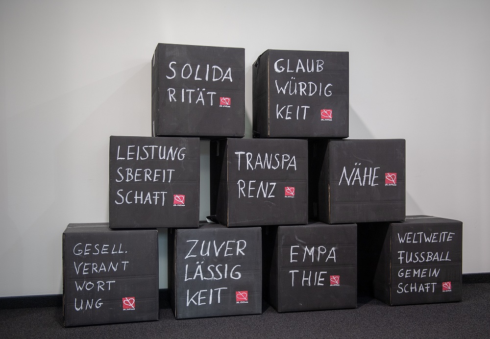 Eine Ansammlung von schwarzen Holzkisten mit weißen Schriftzügen, die verschiedene Werte und Konzepte wie "Solidarität", "Leistungsbereitschaft", "Transparenz" und "Empathie" darstellen. Die Kisten stehen vor einem weißen Hintergrund und symbolisieren die Werte der DFL Stiftung im Rahmen eines Besuchs einer englischen Delegation.