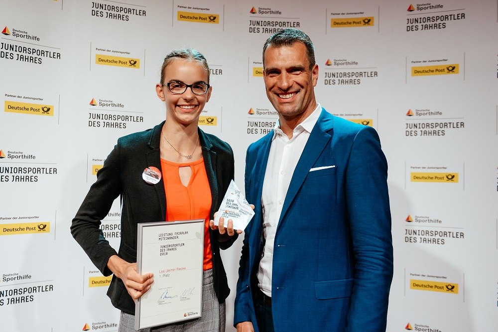 Lea-Jasmin Riecke, Weitspringerin, hält die Auszeichnung "Juniorsportlerin des Jahres 2018" entgegen. Neben ihr steht ein Mann im Anzug. Im Hintergrund sind Banner mit dem Logo "Deutsche Sporthilfe" und der Aufschrift "Juniorsportler des Jahres" zu sehen.