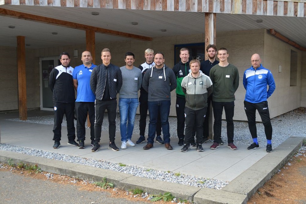 Eine Gruppe von Fußballtrainern steht lächelnd vor einem Gebäude. Sie tragen sportliche Kleidung und sind Teil des Projekts "Jugend - Trainer - STÄRKEN".