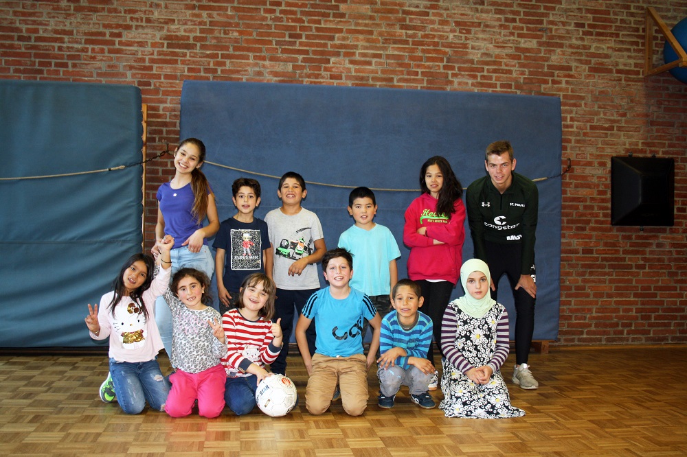 Eine Gruppe von Kindern und Jugendlichen steht in einer Turnhalle und lächelt in die Kamera. Im Hintergrund eine blaue Wand und eine rote Backsteinmauer. Einige der Kinder halten einen Fußball.