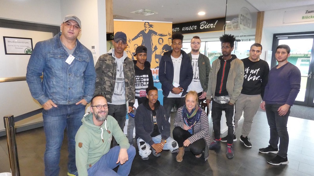 Eine Gruppe junger Männer und Frauen posiert lächelnd für ein Foto. Im Hintergrund ist ein Plakat mit der Aufschrift "Wir lieben unser Bier" zu sehen. Das Bild zeigt Teilnehmer der Job- und Ausbildungsmesse in Fürth im Rahmen des Projekts "Willkommen im Fußball".