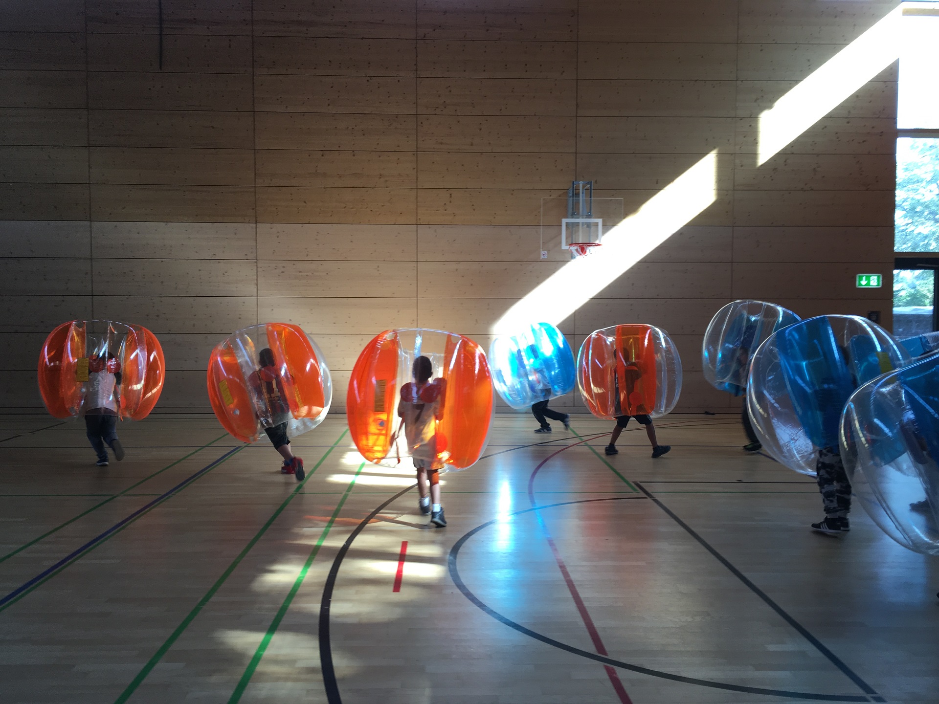 Kinder in aufblasbaren Blasen, sogenannte Bubble Balls, spielen in einer Turnhalle. Im Hintergrund ist ein Basketballkorb und ein Notausgangsschild zu sehen.