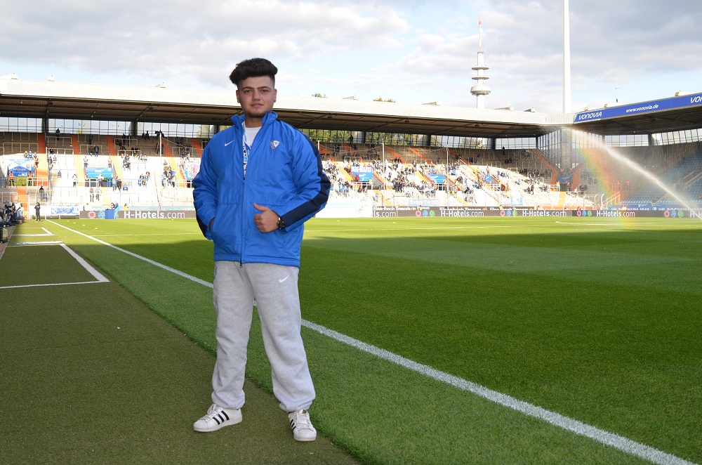 Ein junger Mann steht auf dem Rasen des Fußballstadions. Er trägt eine blaue Trainingsjacke und graue Trainingshose und schaut in die Kamera. Im Hintergrund sind die leeren Zuschauerränge zu sehen.