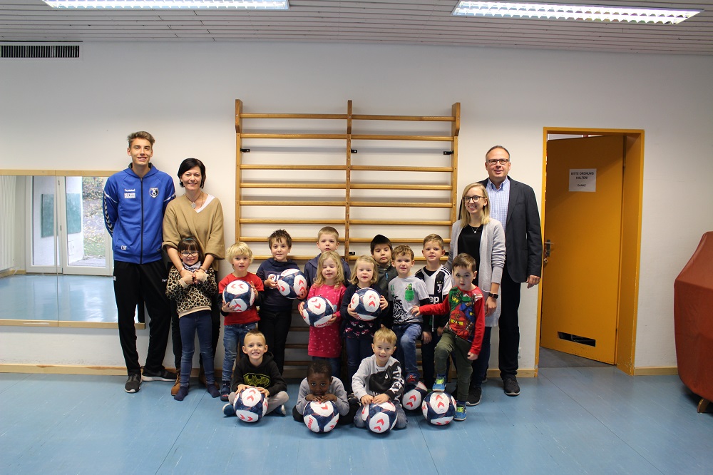 Eine Gruppe von Kindern im Kindergartenalter posiert mit Fußbällen in einer Turnhalle. Neben den Kindern stehen zwei Frauen und ein Mann, vermutlich Vertreter der DFL Stiftung und des Sportclub Lahr. Im Hintergrund ist eine Turnwand und eine Tür zu sehen.