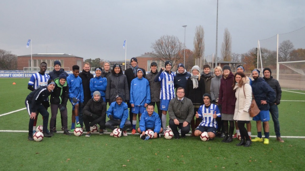 Eine Gruppe von rund 30 Personen steht auf einem Fußballplatz und lächelt in die Kamera. Sie tragen unterschiedliche Kleidung, darunter Trainingsanzüge und Jacken in den Vereinsfarben von Hertha BSC (Blau und Weiß). Einige halten Fußbälle in den Händen. Im Hintergrund ist ein Fußballstadion und kahle Bäume zu sehen.