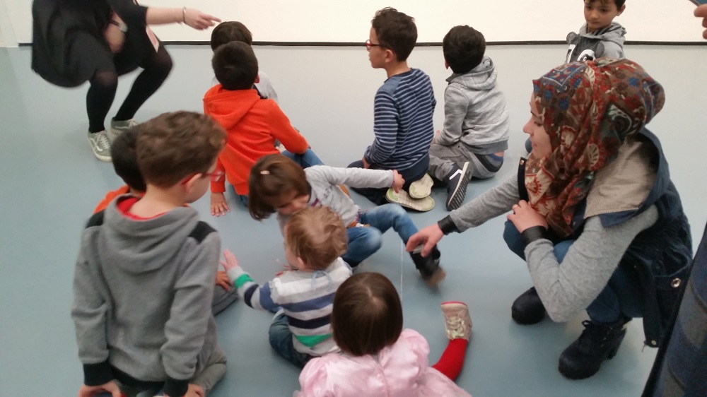 Eine Gruppe von Kindern sitzt im Kreis auf dem Boden und nimmt an einer spielerischen Aktivität teil. Eine Frau mit Kopftuch hockt mit im Kreis, eine andere steht seitlich und zeigt auf etwas. Das Bild zeigt eine Szene aus dem "Willkommen im Fußball"-Bündnis Düsseldorf der DFL Stiftung.