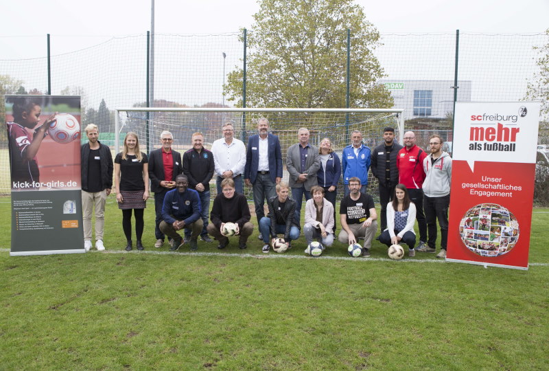 Eine Gruppe von rund 20 Personen steht auf einem Fußballplatz vor einem Tor. Sie lächeln in die Kamera und bilden eine vielfältige Gruppe. Im Hintergrund sind Banner mit den Aufschriften "kickforgirls.de" und "scfreiburg - mehr als Fußball" zu sehen. Das Bild illustriert das "Fußball-Bündnis Freiburg" und das innovative Fortbildungskonzept "Sportlich Sprache lernen".