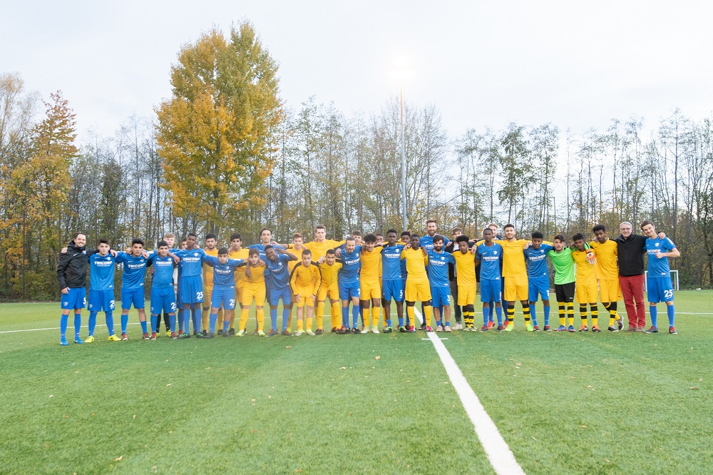 Eine große Gruppe von Jugendlichen steht auf einem Fußballfeld in ihren Trainingsanzügen und lächelt in die Kamera. Einige tragen blaue, andere gelbe Trainingsanzüge. Im Hintergrund sind Bäume mit Herbstlaub zu sehen.