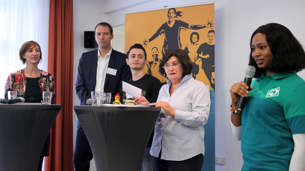 Eine Frau hält auf einem Podium eine Rede, umgeben von weiteren Personen. Im Hintergrund befindet sich ein Banner mit Fußballspielern.