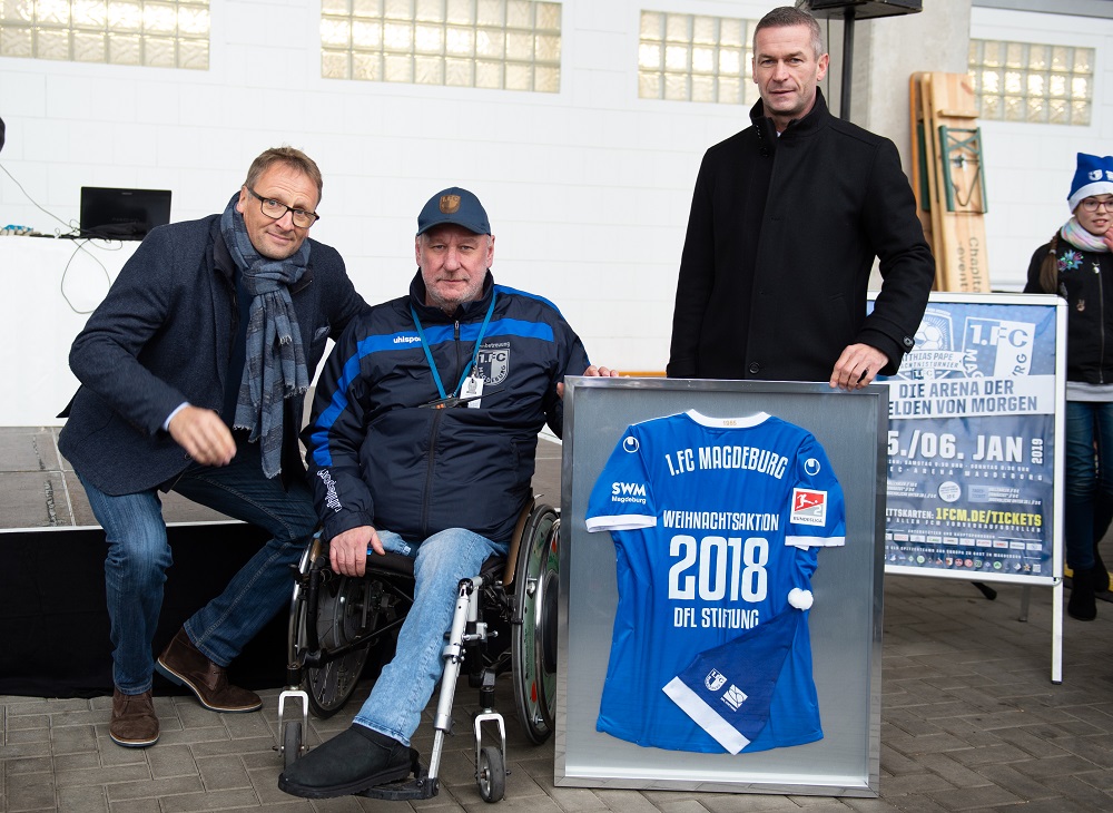 Ein Mann im Rollstuhl nimmt ein blaues Trikot des 1. FC Magdeburg von einem Mann in blauem Jackett entgegen. Im Hintergrund steht ein Werbeaufsteller für eine Veranstaltung des 1. FC Magdeburg.