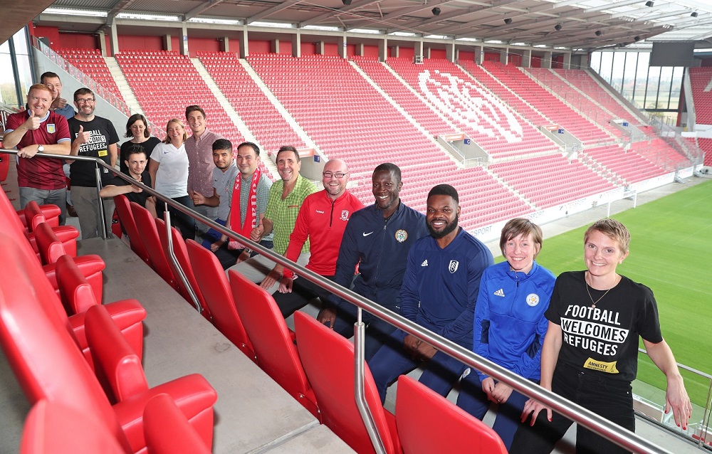 Eine Gruppe von etwa 15 Personen steht auf den roten Sitzen einer Fußballstadiontribüne. Im Vordergrund stehen drei Männer, darunter ein Mann mit dunkler Hautfarbe in Trainingskleidung und ein weiterer in einem blauen Pullover. Hinter ihnen stehen weitere Personen, die die Tribüne füllen. Im Hintergrund ist das grüne Fußballfeld zu sehen.