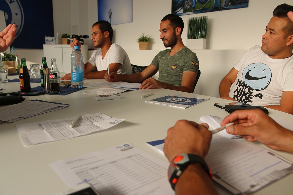 Teilnehmer eines Berufsorientierungskurses sitzen an einem Tisch und schreiben Notizen. Im Hintergrund sind weitere Teilnehmer zu sehen.