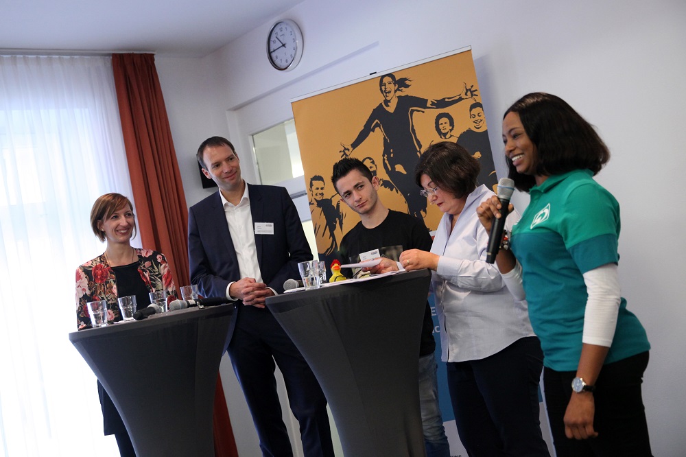 Fachtag "Willkommen im Fußball": Eine Frau hält ein Mikrofon und spricht vor einem Tisch mit mehreren Personen. Im Hintergrund ein Banner mit schematischen Fußballspielern und -fans.