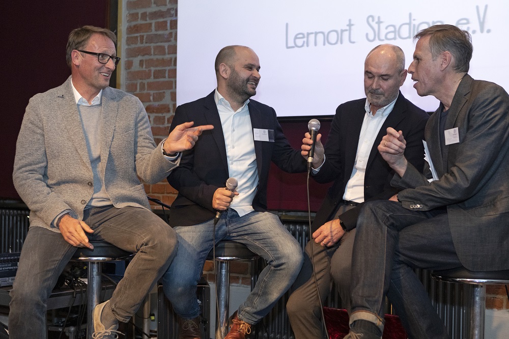 Vier Männer sitzen auf Barhocker auf einer Bühne und diskutieren lebhaft. Im Hintergrund ist ein Banner mit dem Schriftzug "Lernort Stadion e.V." zu sehen. Es handelt sich um eine Veranstaltung des Projekts "Lernen mit Kick" in München.