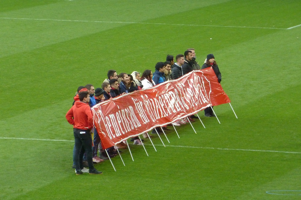 Eine Gruppe von Menschen steht auf dem Rasen eines Fußballstadions und hält ein großes Banner mit der Aufschrift "ERINNERN. WIEDERERLEBEN. BEWAHREN." hoch. Im Hintergrund ist grünes Spielfeld zu sehen.