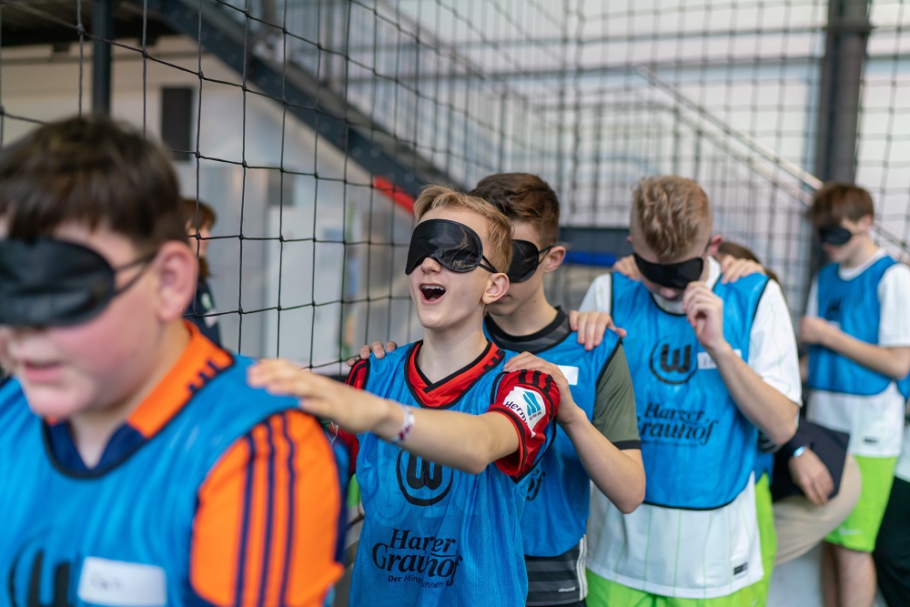 Eine Gruppe Jugendlicher mit Augenbinden nimmt an einem Fußballworkshop im Rahmen der Paralympischen Woche der DFL Stiftung teil. Ein Junge im blauen Trikot mit dem Schriftzug "Haro" steht im Vordergrund und scheint mit Armen und Beinen in Bewegung zu sein. Im Hintergrund sind weitere Teilnehmer im gleichen Trikot zu sehen.