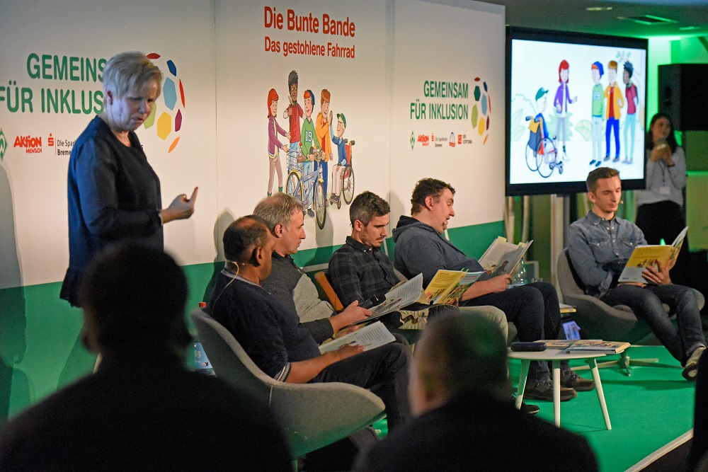 Fußballspieler und Unterstützer sitzen auf einer grünen Bank in einem Raum und lauschen einem Vorleser. Im Hintergrund eine Wand mit dem Logo der Aktion Mensch, des SV Werder Bremen und der DFL Stiftung sowie der Aufschrift "Gemeinsam für Inklusion" und einer Illustration von Menschen mit und ohne Behinderung auf Fahrrädern.