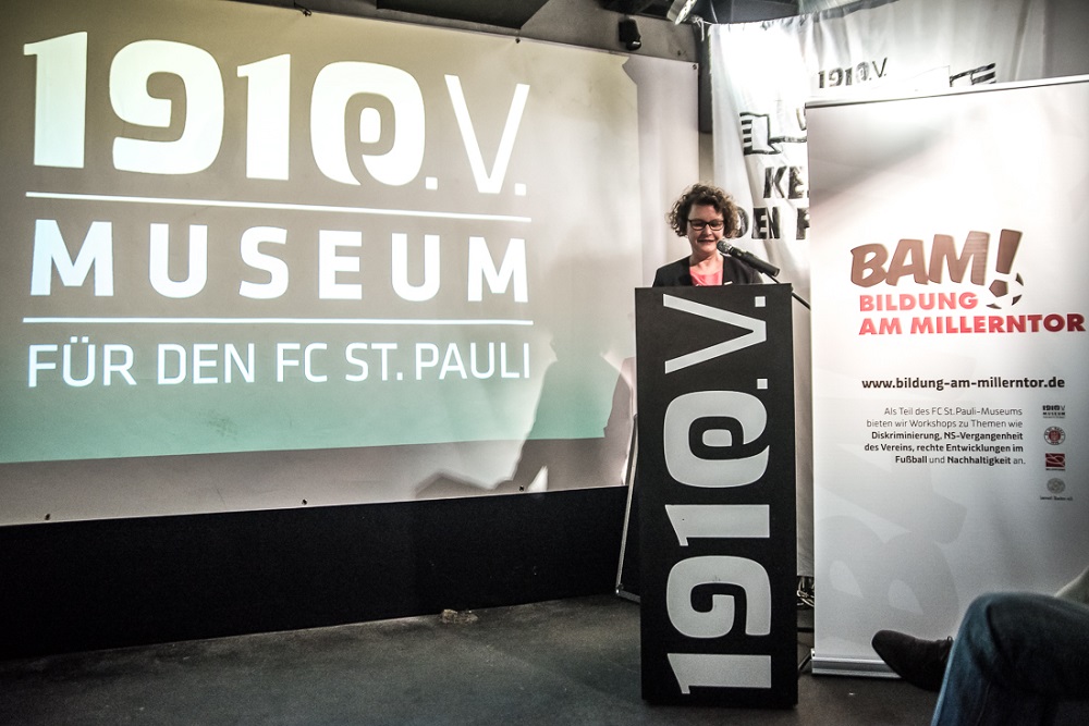 Eine Frau steht vor einer Wand mit dem Aufdruck "1910V MUSEUM FÜR DEN FC ST. PAULI" und dem Logo "BAM! Bildung am Millerntor". Sie spricht in ein Mikrofon und hält eine Rede zur Eröffnung des 20. Lernorts Stadion beim FC St. Pauli.