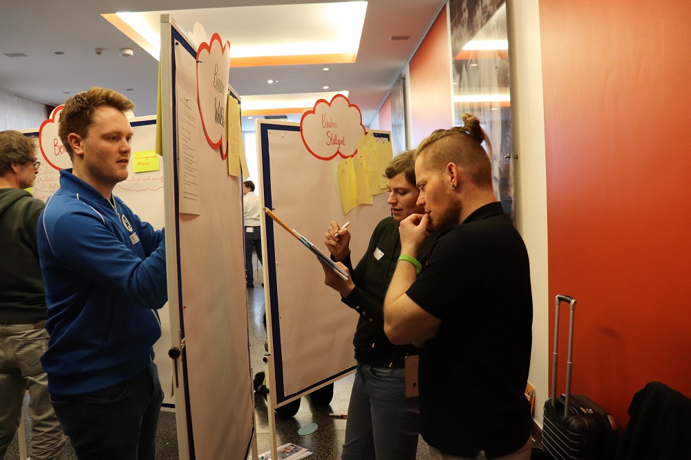 Eine Gruppe von Menschen steht in einem hellen Raum vor Flipcharts, die mit Text und Zeichnungen beschriftet sind. Sie betrachten die Inhalte der Charts aufmerksam während eines Auftakttreffens der DFL Stiftung.