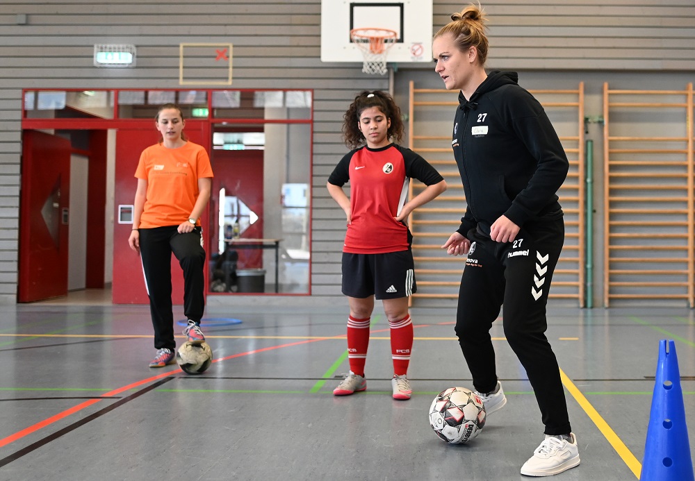 Clara Schöne und Hasret Kayikci der DFL Stiftung führen eine Fußball-AG in der Emil-Thoma-Schule durch. Im Bild sind die beiden Trainerinnen mit jungen Mädchen in einer Sporthalle zu sehen, während sie Fußballtechniken üben.